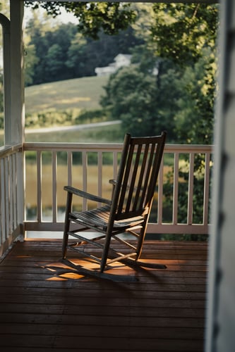 A weathered wooden rocking chair on a covered porch with white railings, bathed in warm golden-hour sunlight, overlooking green rolling hills and trees.