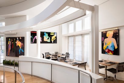 A modern restaurant dining area with sleek wooden tables and glassware set for service. Large windows allow natural light, and vibrant artwork decorates the walls.