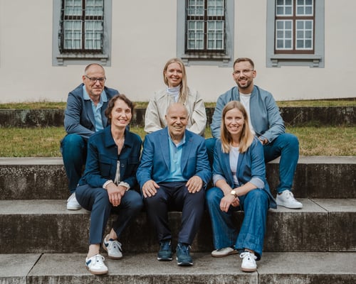 Das Team der Deutschen Fördermittelberatung lächelt freundlich auf Steinstufen vor einem historischen Gebäude. Die sechs Fördermittelberater tragen harmonisch abgestimmte Kleidung in Blau- und Beigetönen und vermitteln eine kompetente sowie vertrauenswürdige Atmosphäre.
