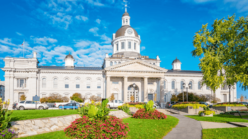 Historic building with a large dome and clock tower under a bright blue sky, surrounded by manicured gardens with colorful flowers and green lawns.