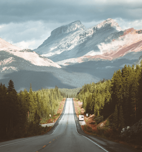 A long, empty highway stretches into the distance, flanked by dense green forests. Majestic mountains loom under a cloudy sky, creating a serene landscape.