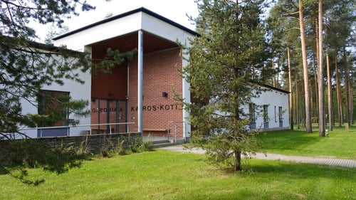 Julkisivukuva Kairos-kodista. Rakennus on valkoinen, mutta sisäänkäynnissä on punatiilistä seinää ovien vieressä. Aivan rakennuksen vieressä on viheraluetta ja metsää, jossa kasvaa havupuita.