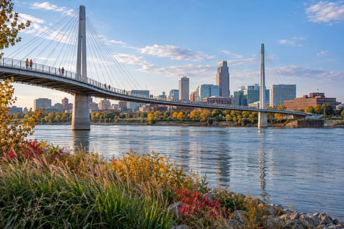 Bob Kerrey Pedestrian Bridge spanning the Missouri River, with downtown Omaha’s skyline, colorful autumn foliage, and a blue sky.