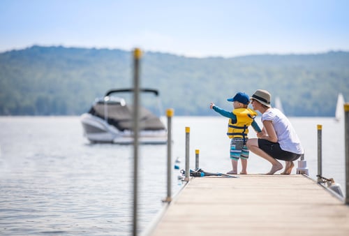 A child in a yellow life jacket and blue cap stands on a wooden dock pointing at a boat, with an adult beside them. A calm lake and hills in the background.