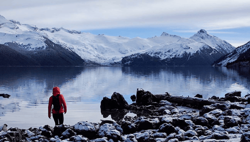 A person standing along a rocky shoreline, with a calm lake and snow-peaked mountains in the background.