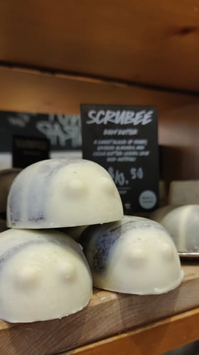 Unpackaged shower scrub in the shelf of a bee sits on a wooden shelf. Sign next to it reads Scrubee.