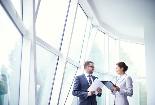 Two professionals in formal attire are talking near modern, sunlit windows. The man holds documents, and the woman holds a clipboard, indicating discussion.