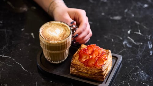 A woman's hand reaches for a foamy heart-topped mocha that sits next to a freshly baked pastry with strawberries at Tasse Coffee Shop at the Four Seasons Chicago.