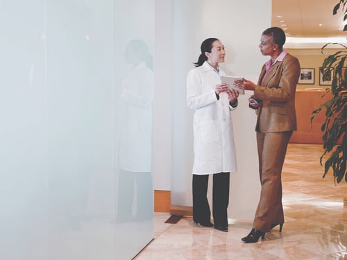 A doctor in a white coat and a businesswoman in a brown suit are engaged in a conversation in a bright, modern office. The mood is professional and collaborative.