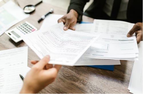 Two individuals exchanging documents over a desk with paperwork, a pen, and a calculator in view. The scene conveys a professional, collaborative atmosphere.