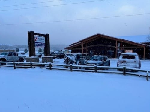 A snow-covered Builders FirstSource location with several vehicles parked in front of a rustic, wood-frame building. Snow is actively falling, covering the ground, roof, and vehicles. A large Builders FirstSource sign stands near the entrance, and a wooden fence lines the snowy foreground.