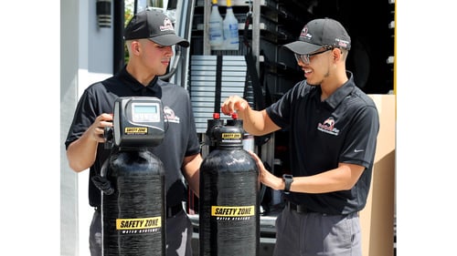 Two First Choice Plus plumbers preparing Safety Zone whole-home water-treatment tanks beside a service vehicle.