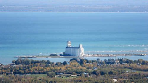 Aerial view of a large, white grain elevator and silos by a calm lake. Foreground features lush green trees and scattered residential homes.