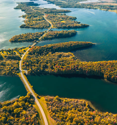 Aerial view of a winding road crossing narrow forested islands, surrounded by blue water, with trees in autumn colors. The scene is serene and picturesque.