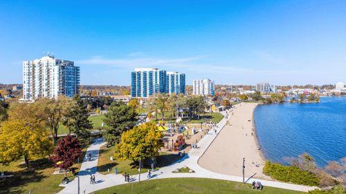 Aerial view of a vibrant Barrie Ontario beachfront with sand, blue water, and a nearby park. People stroll on paths, surrounded by colorful autumn trees and modern buildings.
