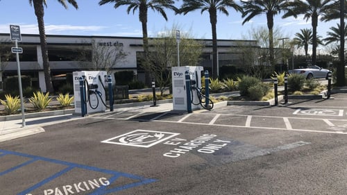 Several EVgo fast charging units installed in marked parking spaces at a public charging station, with EVgo logos on the chargers, protective bollards around each unit.
