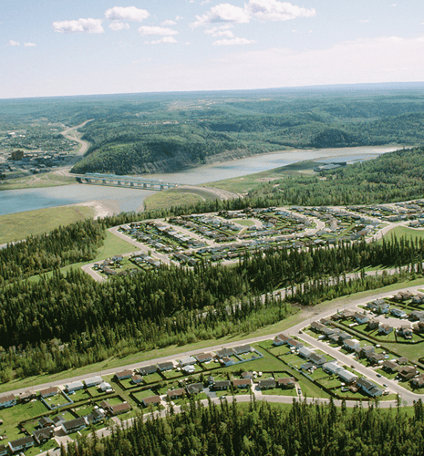 Aerial view of a suburban neighborhood with neatly arranged houses, surrounded by lush green forests. A river and bridge stretch in the distance under a bright, clear sky.