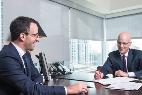 Two male professionals in navy suits sitting across from each other at a dark wooden desk in a modern office with floor-to-ceiling windows overlooking a city skyline. They appear to be in a meeting or consultation, with documents on the desk between them. The office features contemporary furnishings including a desk lamp and office equipment, creating a polished, corporate atmosphere.