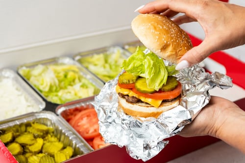A person places the top bun on their cheeseburger. A Five Guys catering box full of toppings is in the background.