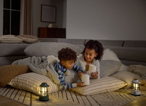 Two kids sip hot cocoa while laying on the floor in their living room. The area is lit with lanterns.