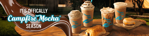 An assortment of iced and blended Caribou Coffee drinks with whipped cream on a wooden table, alongside a breakfast sandwich and croissant.