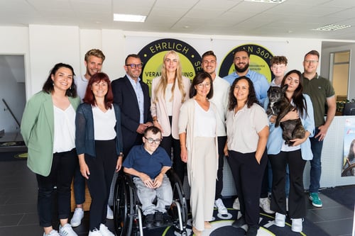 Gruppenfoto von dreizehn Personen in einem Büro. Eine Person sitzt im Rollstuhl, eine andere hält einen kleinen Hund im Arm. Die Gruppe wirkt freundlich und divers, mit Personen unterschiedlichen Alters und Erscheinungsbildes. Im Hintergrund ein Firmenlogo mit den Worten „EHRLICH“ und „VERANTWORTUNG“.