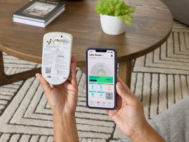 A person holding a smart Kidde CO alarm next to their phone, showing the Kidde Advanced Home Health App.