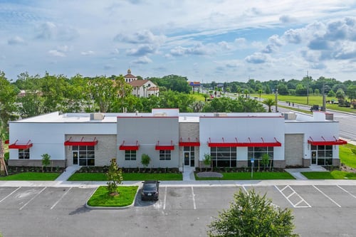 An elevated exterior view of a modern, single-story medical building for the Cardiovascular Institute of Central Florida. The building features a white and gray facade with red awnings over the windows and doors, tan stone accents, and a paved parking lot in the foreground.