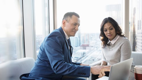 Man in a blue suit and woman in a gray sweater collaborate on a laptop in a modern office with large windows overlooking a cityscape.