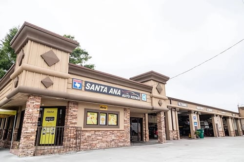 Exterior view of 'Santa Ana Auto Repair with beige architecture and wooden detailing, featuring a large blue sign and adjacent 'Hertz' rental advertisements, alongside multiple covered service bays."