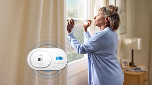 A woman opens her window. In the foreground, a carbon monoxide alarm is going off, showing that 39 ppm has been detected in the air.  Blue circles representing the sound is emanating from the device.