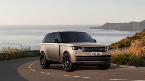 Gold Range Rover driving along a coastal road with hills and ocean in the background on a sunny day.