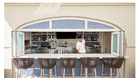 A bartender in a white shirt preparing a poolside bar with blue mosaic tiles, surrounded by four high-back wooden chairs. Bottles and glasses are visible on shelves behind him.