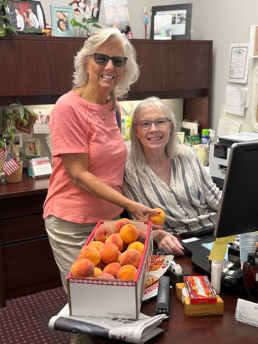 Two women sitting at a desk with a box of peaches in front of them.