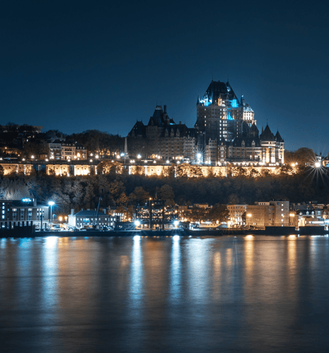 A stunning night view of a city skyline with a prominent, illuminated castle-like building atop a hill, reflecting on the calm water below.