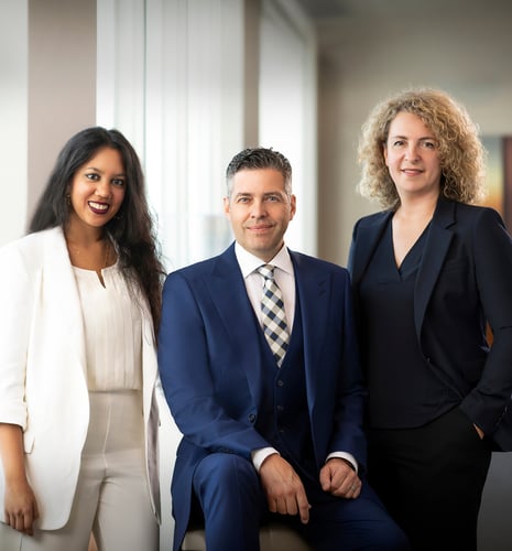 Three professionals, two women and one man, pose confidently in business attire. The man is seated in the center wearing a blue suit, flanked by the women standing in white and black suits, suggesting teamwork and professionalism.