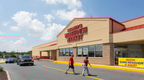 Black car driving towards two people in red shirts walking across parking lot to Redner's Warehouse Market at Whitehall Square shopping center