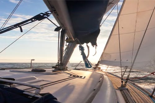 Sailboat deck with billowing sails, ropes, and calm ocean horizon at sunset.