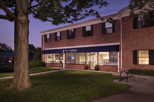 Two-story brick building with black shutters housing William Pitt Sotheby's International Realty in Ridgefield, Connecticut, photographed at dusk with illuminated storefront windows.
