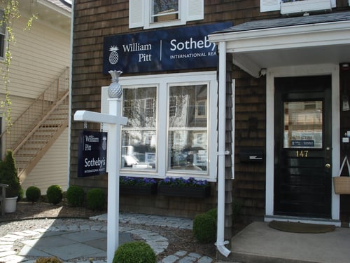 Brown shingled building with white trim housing William Pitt Sotheby's International Realty in Rowayton, Connecticut, featuring decorative pineapple post and window flower boxes.