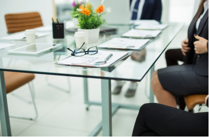 Glass conference table with documents, glasses, and a pen. People in business attire sit around it; a plant centerpiece adds color to the modern office setting.