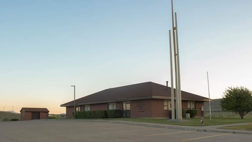 The outside of a church building. There is a separate steeple on the corner of the building. A parking lot with automobiles, grass, and a large pine tree surround the church.