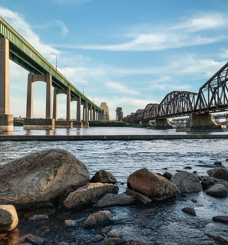 Two bridges span a river under a blue sky. The left bridge is modern with tall pillars, while the right is a rustic metal truss. Rocks line the riverbank.