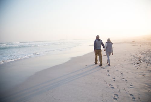 An elderly couple walks hand in hand along a serene beach at sunrise, casting elongated shadows on the sand. Waves gently lap the shore, creating a peaceful atmosphere.