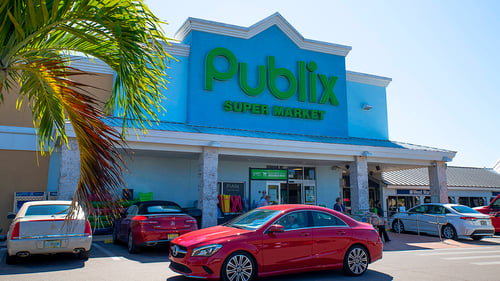 Red car driving through parking lot of Publix with a palm tree on the left