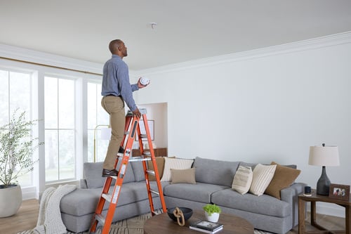 A man on a ladder is installing a new smoke alarm.