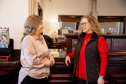 Two females engaged in a friendly conversation at a Bryant Bank teller counter. One wears a taupe blouse while the other wears a red sweater and a black quilted vest. The background shows a warm, professional office environment with dark wood cabinetry and a lamp.