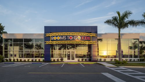 Exterior of Rooms To Go Kids store in Fort Myers, Florida, with a blue facade and large windows reflecting the sunset. The "Rooms To Go Kids" sign is prominently displayed above the entrance.
