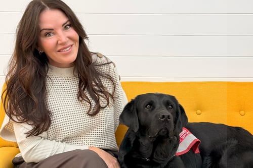 Rhonda Hymers and service dog sit together on bright yellow couch against white wall.