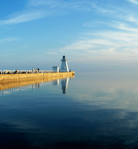 A white lighthouse with a green top stands on a pier, reflecting in the calm water. A few people walk along the pier under a clear blue sky.